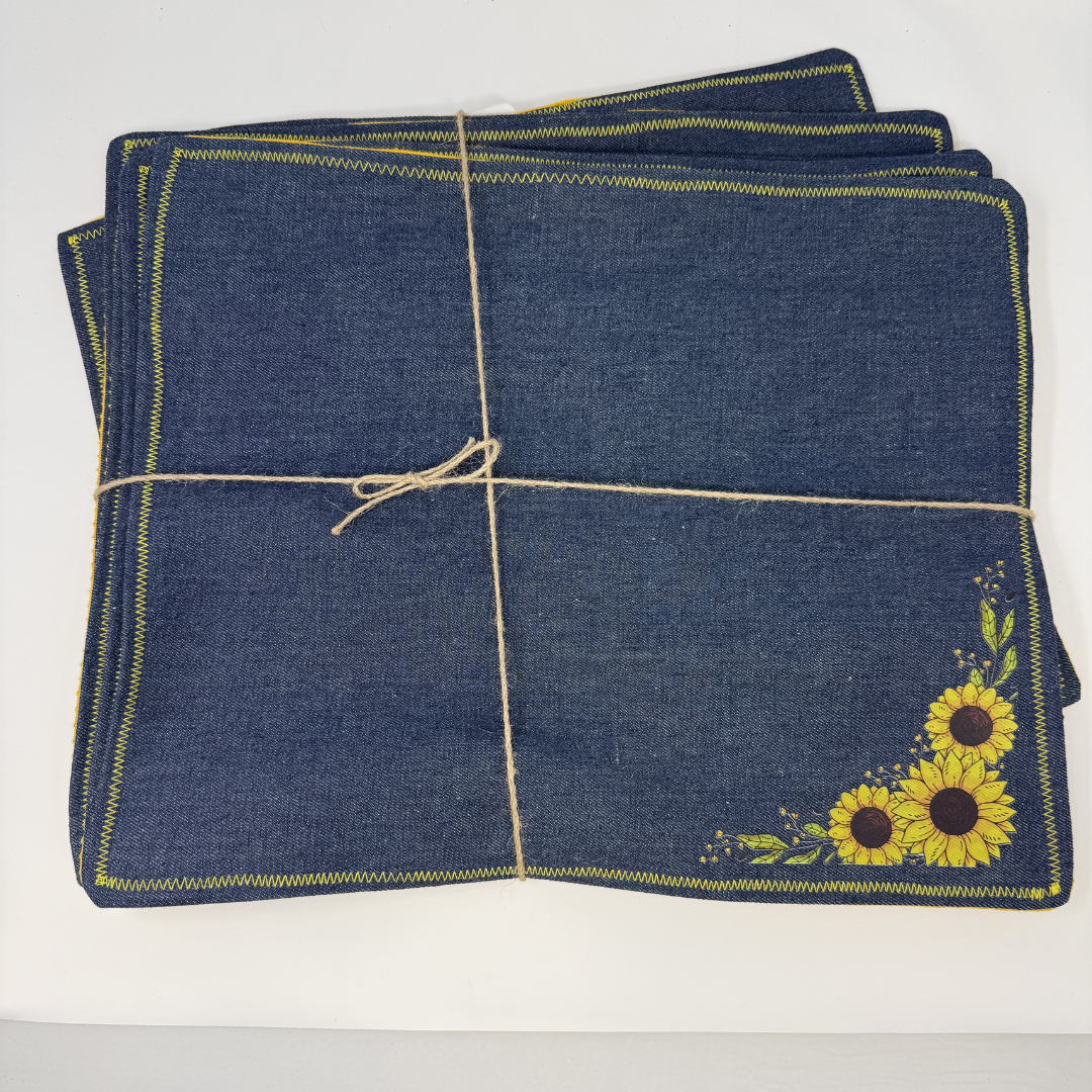 A stack of dark blue denim placemats tied with twine, featuring yellow and brown sunflower embroidery in the bottom right corner and yellow stitched borders.